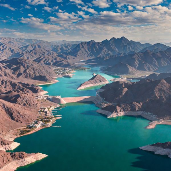 Aerial drone panoramic view of vibrant turquoise Hatta Dam reservoir in Dubai UAE, surrounded by rugged reddish-brown Hajar Mountains at golden hour sunset